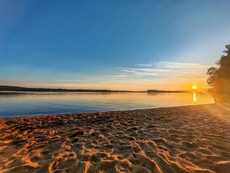 Serene sunset over a calm lake with a sandy beach in the foreground, reflecting golden and orange hues in the water.