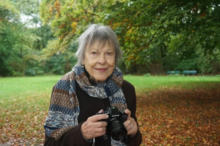 Older woman holding a camera, smiling warmly, with a scenic outdoor background.