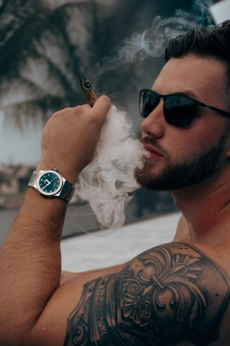 Young man with a tattooed bicep holding a cigar, his confident gaze directed forward, set against a neutral background.